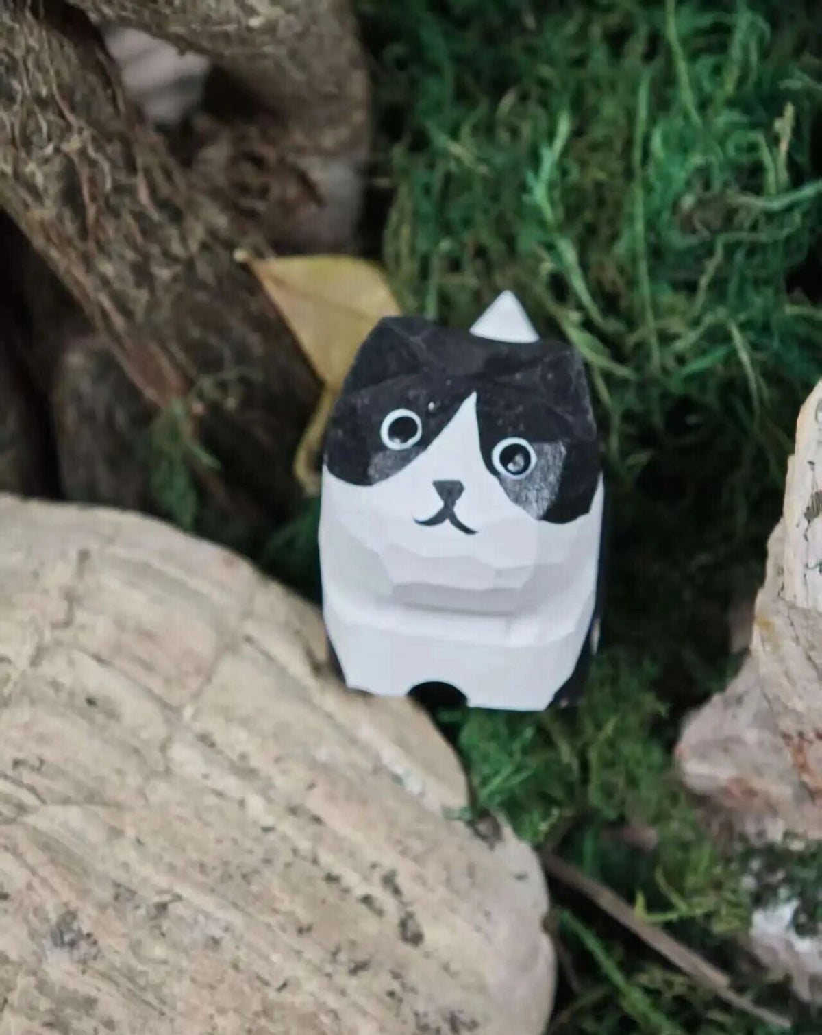 A small black and white cat-shaped paper toy or figurine placed among moss, rocks, and branches in an outdoor-style setting.