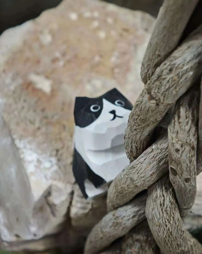 A close-up of a small black-and-white paper cat figurine nestled among rough driftwood branches and pale stones.