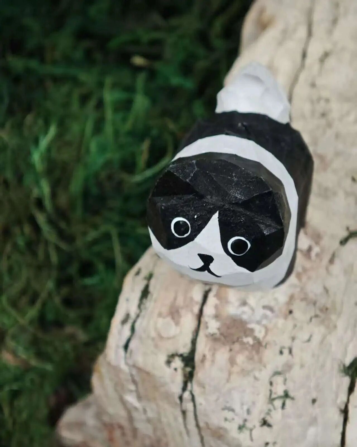 A hand-painted wooden tiger figurine with a smiling face, resting on a stone next to green moss and natural wood pieces.