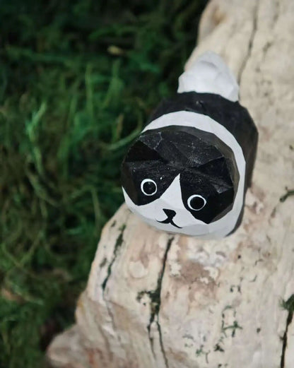 A hand-painted wooden tiger figurine with a smiling face, resting on a stone next to green moss and natural wood pieces.