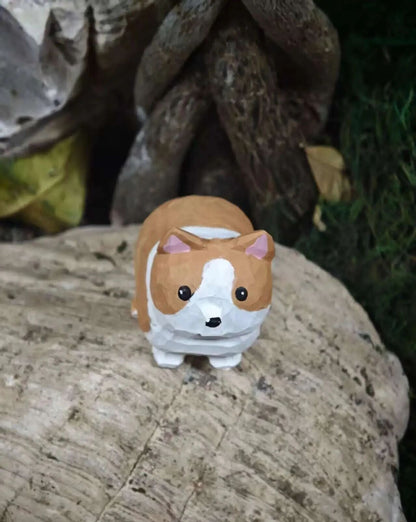 A small painted corgi figurine standing on a textured log outdoors, with blurred tree roots and greenery in the background.