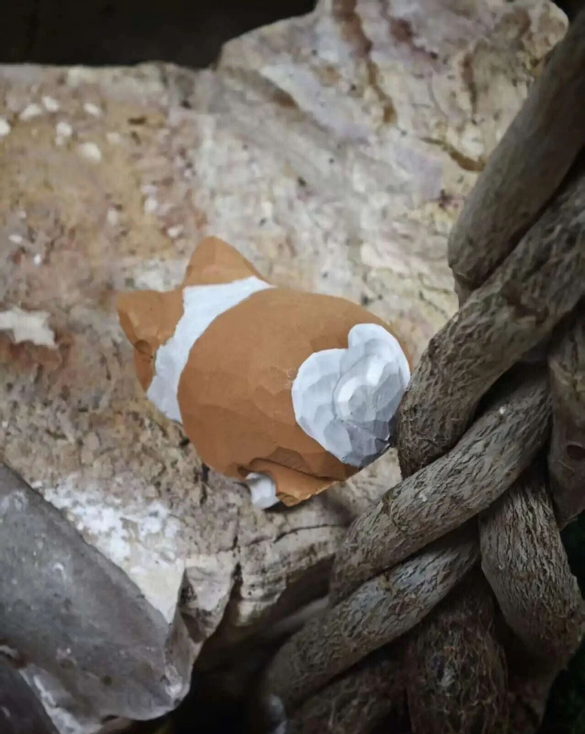 A small sculpted figurine resembling a cat or animal with orange-brown, white, and gray sections, placed among rough rocks and twisted branches in a natural-looking setting.