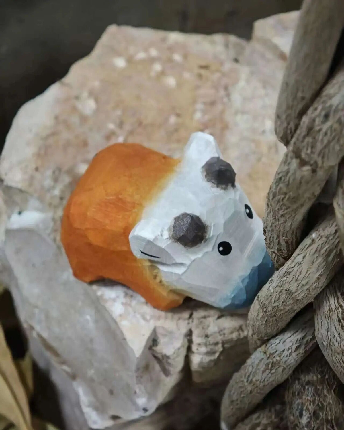 A small hand-painted wooden animal figurine, resembling a stylized bear or raccoon, resting on a textured stone next to a rustic wooden post.