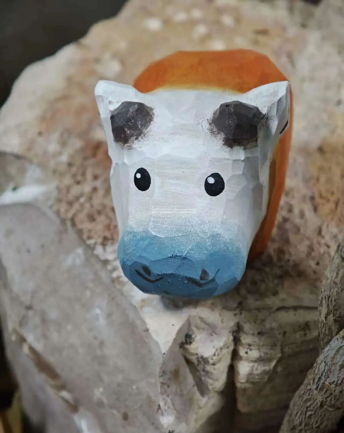 A small hand-painted wooden cow figurine with a white face, black ears and eyes, a blue snout, and an orange top, displayed on a textured stone surface.