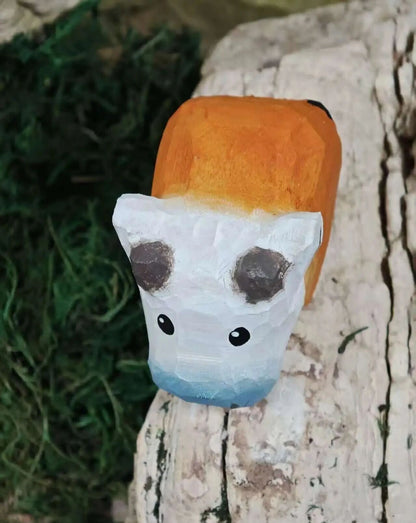A hand-painted wooden cow-shaped lantern or decorative object with an orange top, white face, and blue lower section, resting on a weathered tree trunk outdoors.