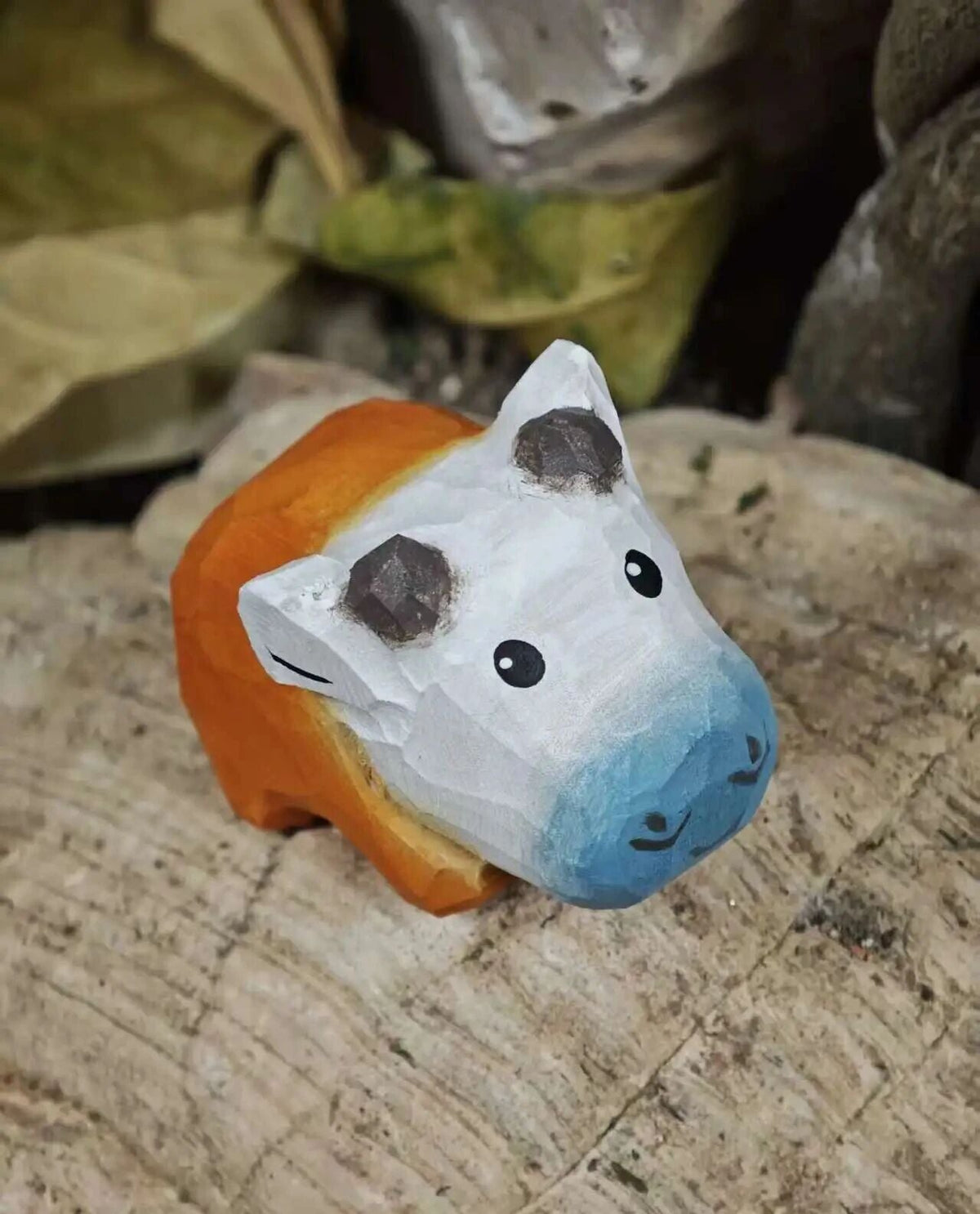 A small hand-painted wooden cow figurine with a white face, blue nose, orange body, and dark brown horns, resting on a textured tree stump.