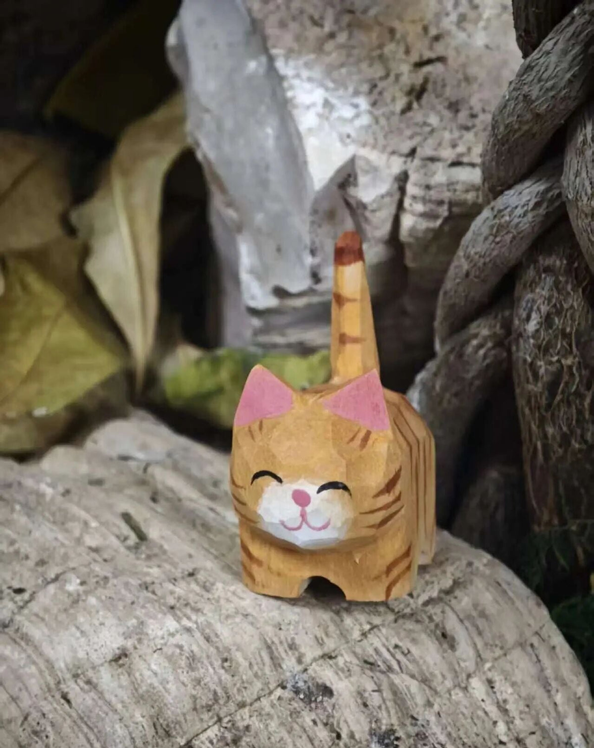 A small hand-carved wooden cat figurine with orange stripes, pink ears, and a white muzzle, standing on a textured stone surface in a rustic outdoor setting.