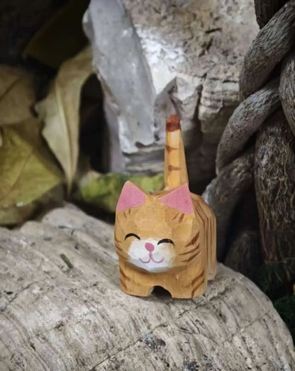A small hand-carved wooden cat figurine with orange stripes, pink ears, and a white muzzle, standing on a textured stone surface in a rustic outdoor setting.