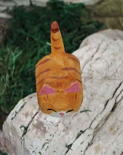 A small orange cat-shaped decorative object with pink inner ears, closed black eyes, and a pink nose, resting on a white textured stone with green foliage in the background.