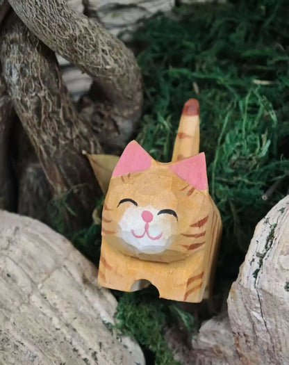 A hand-carved wooden cat figurine with pink ears and a smiling face, nestled among green moss and natural wood logs.