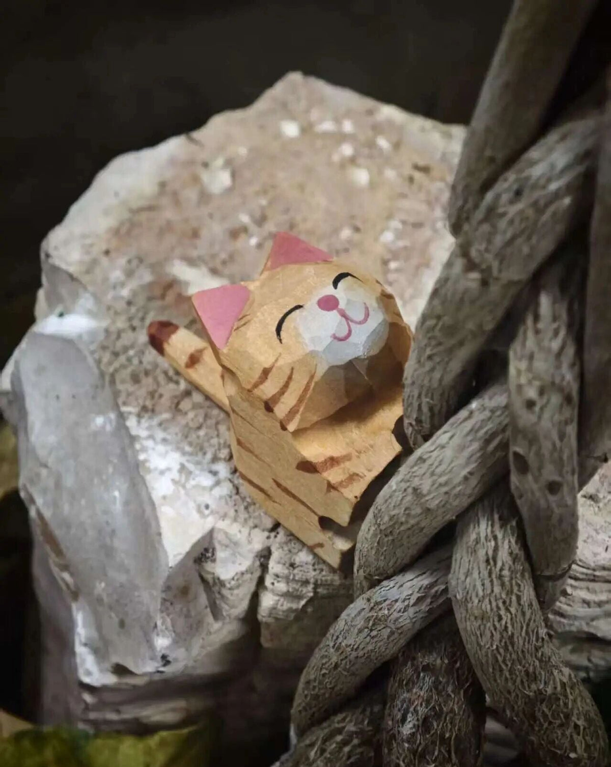 A small wooden cat figurine with a paper-like face and pink ears, resting on a rough stone beside a thick braided rope.