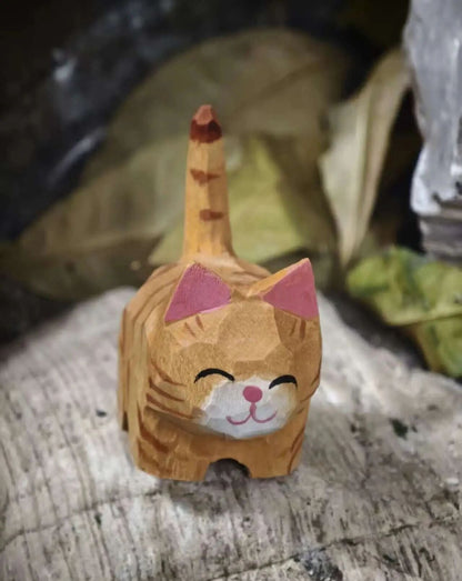 A hand-carved wooden cat figurine with orange-brown stripes, pink ears, and a white muzzle, standing on a weathered wooden surface with a blurred background.