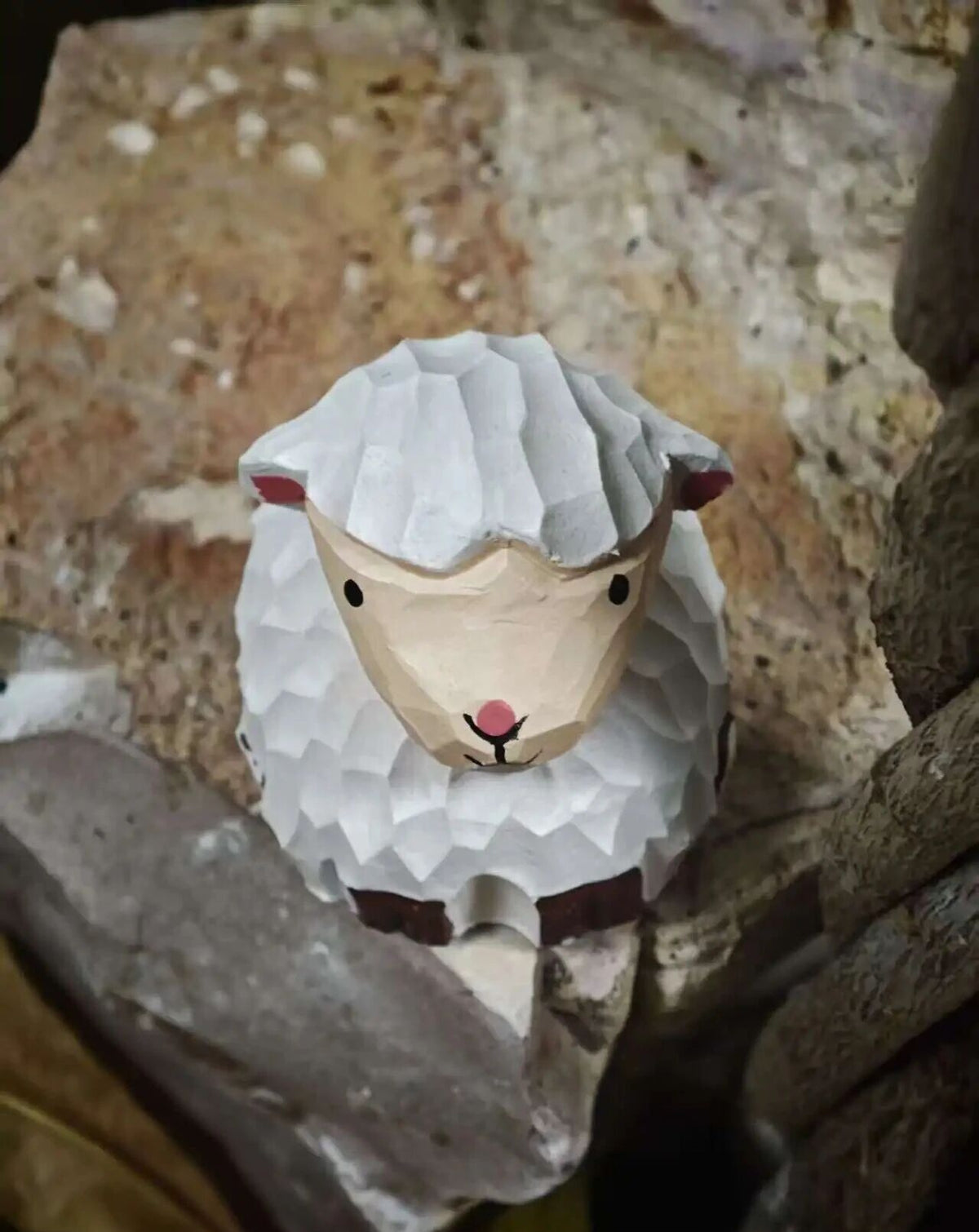 A small handcrafted sheep figurine with a geometric, faceted texture, featuring a cream-colored face, white textured wool, and small pink and black details, set against a rustic stone background.