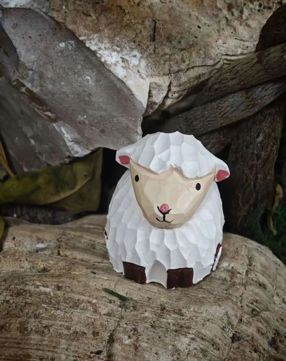 A small hand-painted ceramic sheep figurine with textured white wool, a beige face, pink ears and nose, and dark brown accents, displayed on a rustic wooden log with natural rocks in the background.