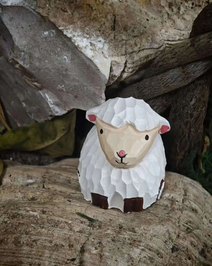 A small hand-painted ceramic sheep figurine with textured white wool, a beige face, pink ears and nose, and dark brown accents, displayed on a rustic wooden log with natural rocks in the background.