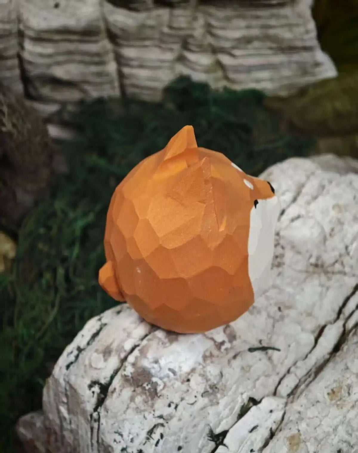 A low-poly geometric fox figurine in orange and white, sitting on a white textured stone surface with moss and layered rocks in the background.
