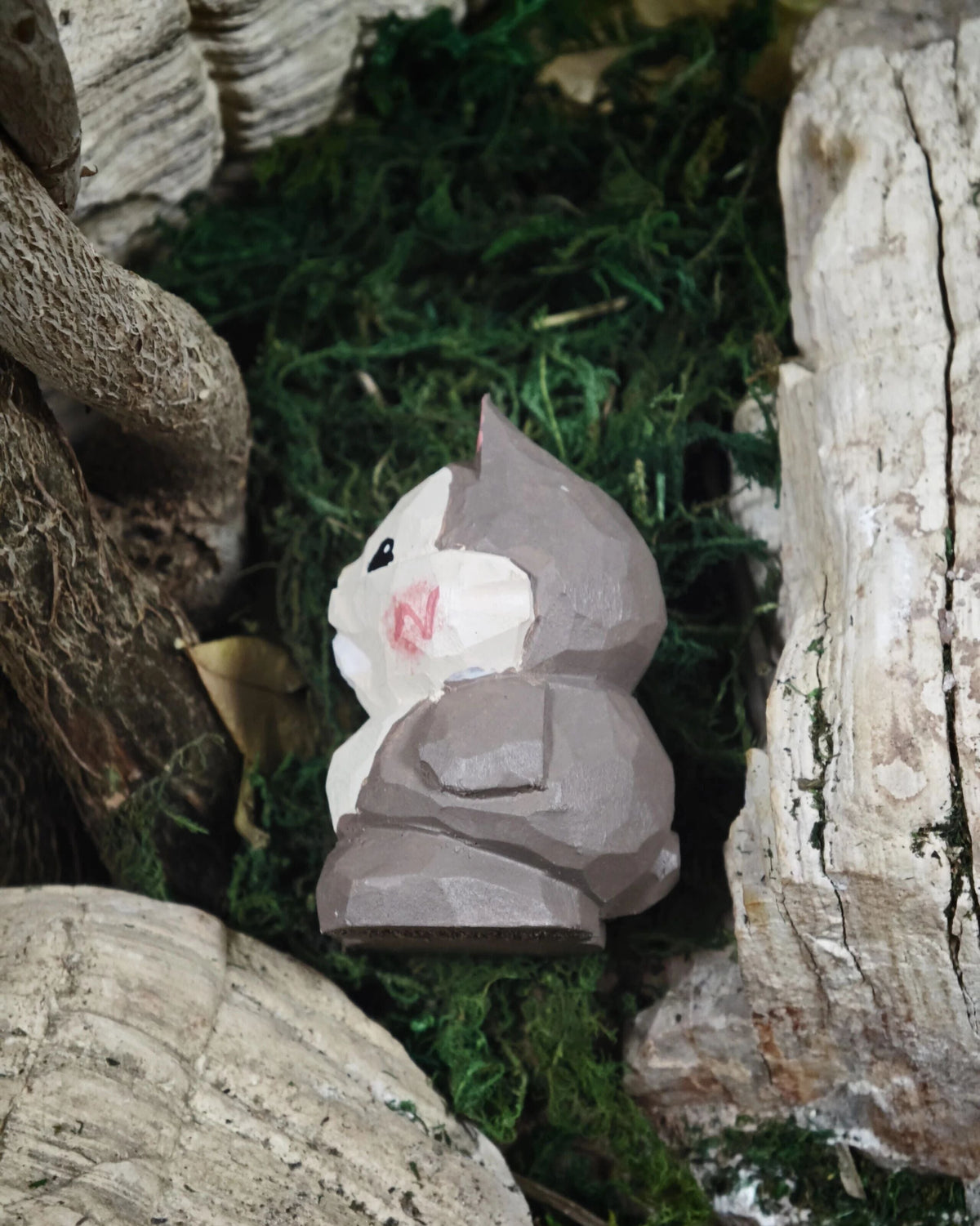 A small hand-carved wooden figurine of a cat wearing a gray cloak, resting on green moss between natural wood logs.