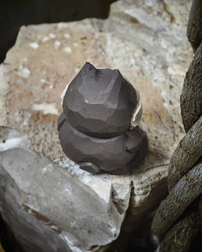 A dark gray, faceted, sculptural cat figurine sitting on rough, natural stone slabs.