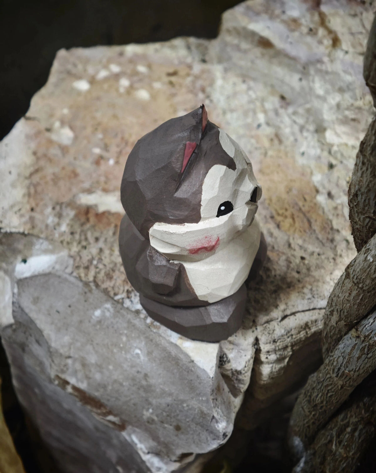 A handmade wooden animal figurine, shaped like a cat or small animal with a geometric, faceted design, resting on rough natural rocks.