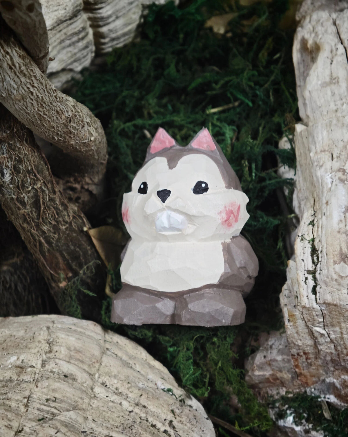 A small painted hamster figurine with pink ears and pink cheeks, sitting on green moss between rustic wood logs.