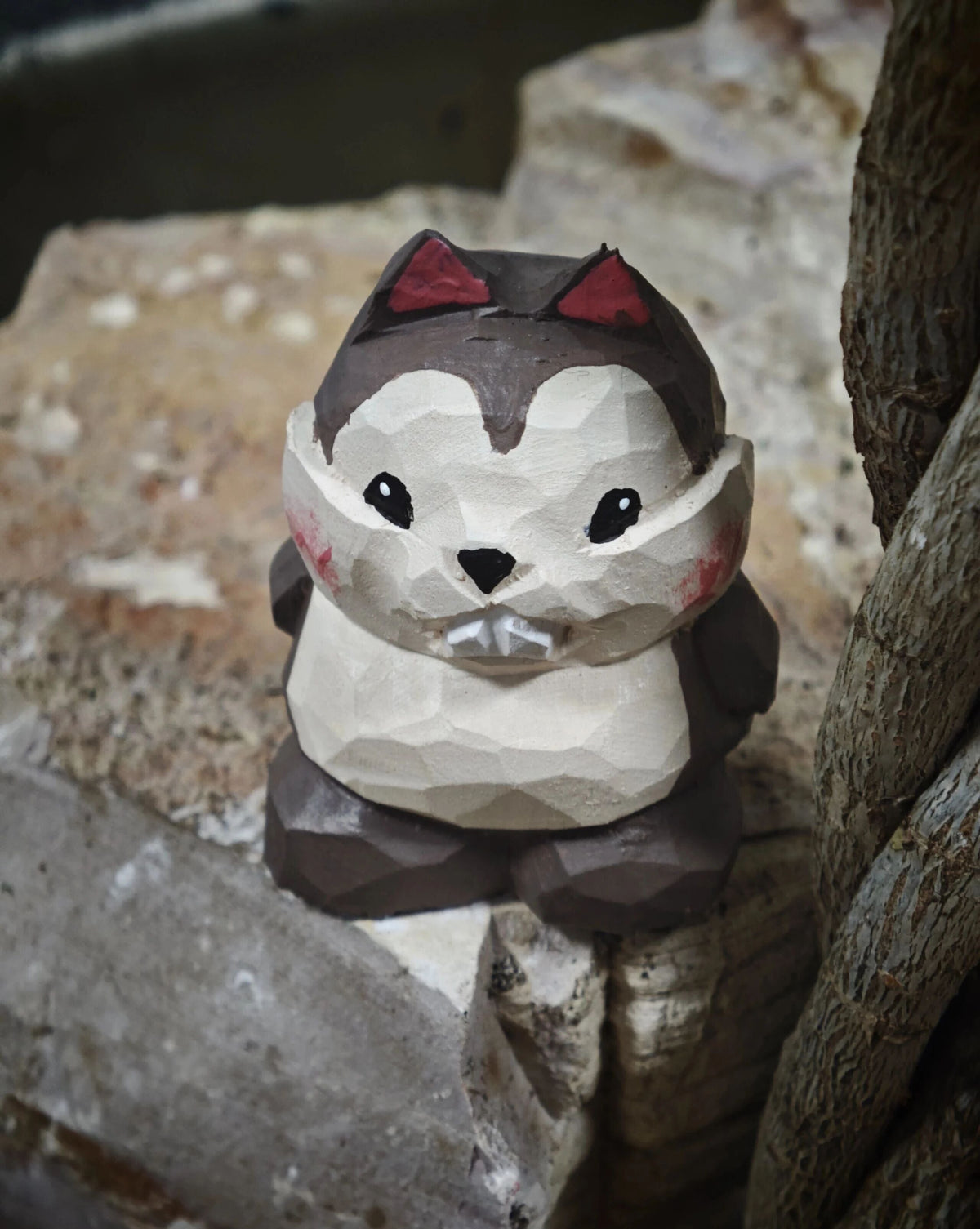 A low-poly style wooden beaver figurine with a dark brown and cream color scheme, red ear accents, and a small white tooth, set against a rustic background of rocks and wood.