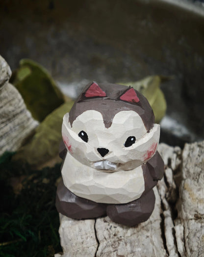 A low-poly wooden animal figurine shaped like a cute brown and white bear or chipmunk with pinkish-red ear accents, sitting on a piece of weathered wood.