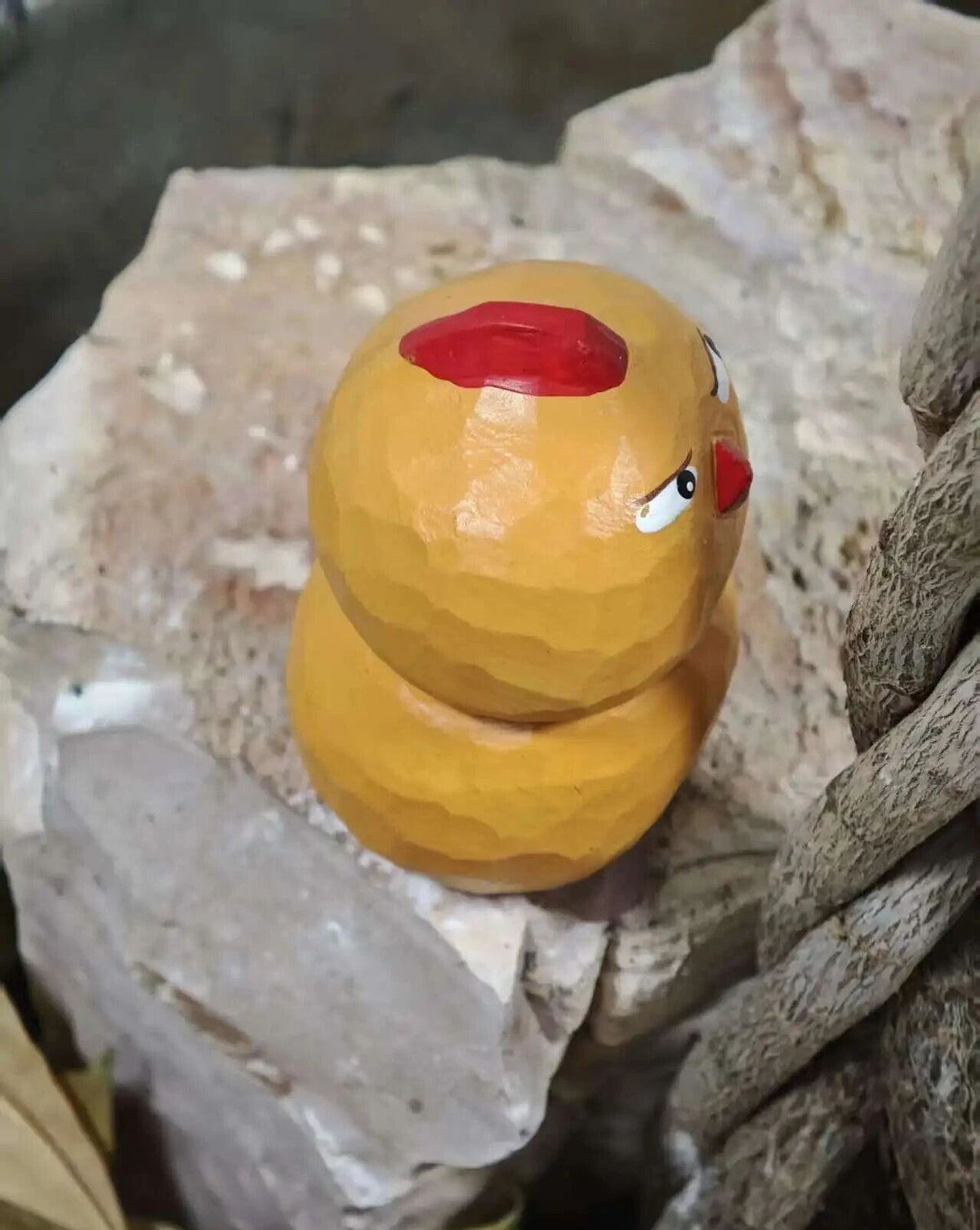 A small handcrafted wooden toy figure resembling a stylized chicken or duck, painted in yellow-orange with a red top detail and a red beak, resting on rough natural stones.