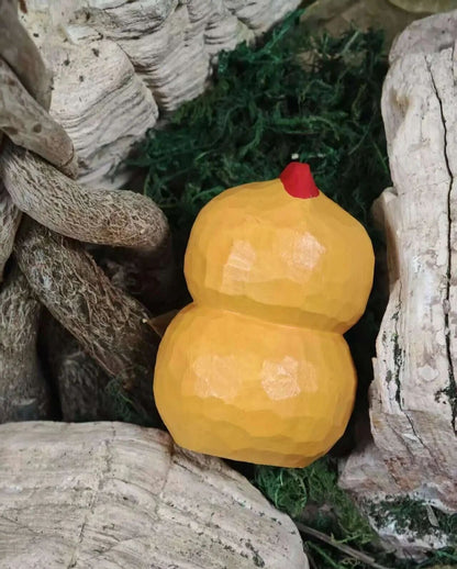 A small decorative double-gourd ornament in a warm golden yellow color with a tiny red top, nestled among faux moss and natural wood driftwood.