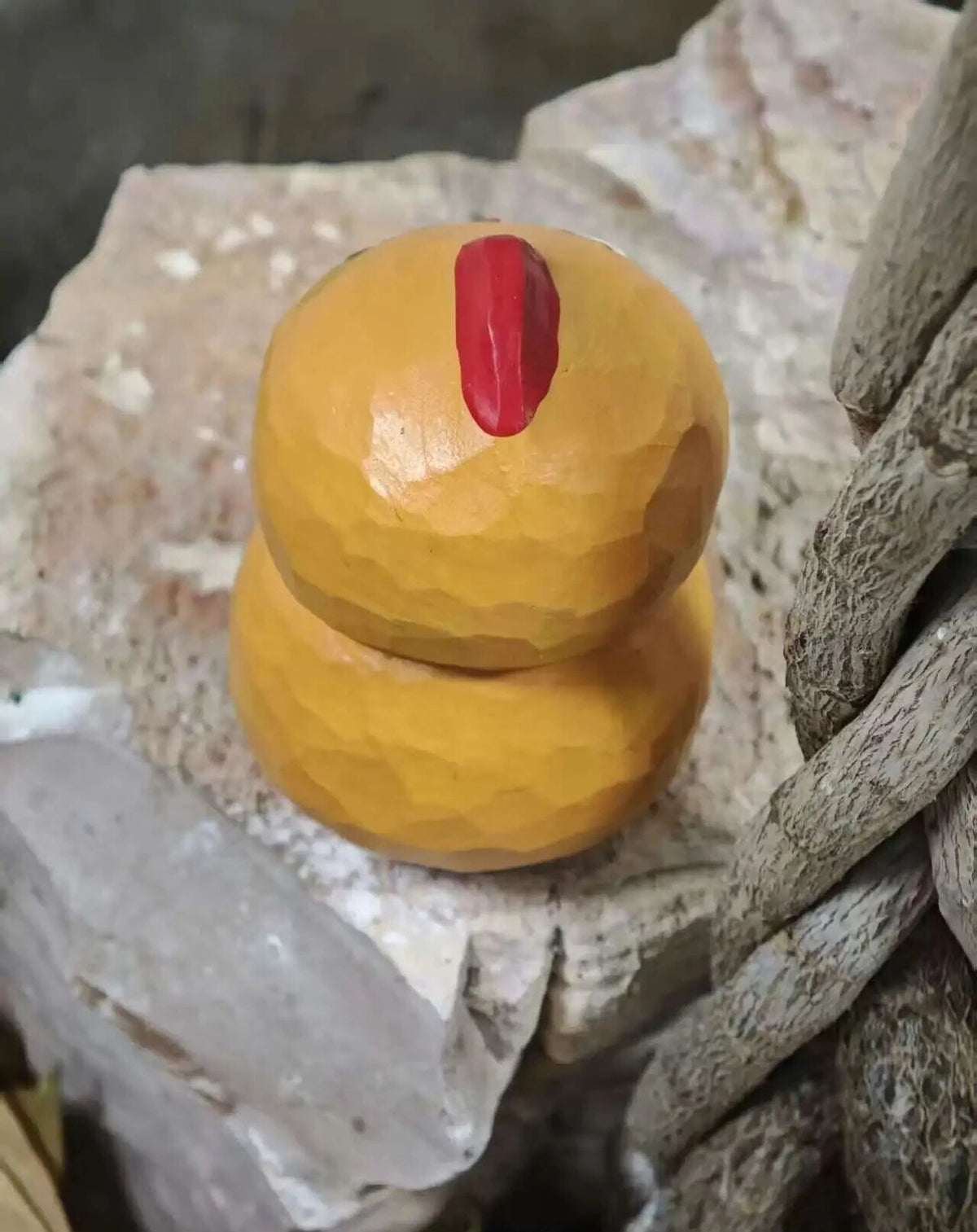A small hand-carved wooden figurine shaped like a stylized chicken, made from two stacked natural gourd-like forms with a red painted beak, displayed on rough stones.