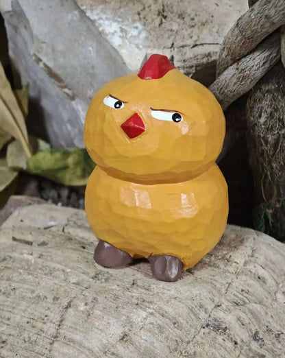 A small hand-carved wooden chicken figurine with a grumpy expression, featuring a yellow-orange body, red comb and beak, and brown feet, standing on a rustic log.