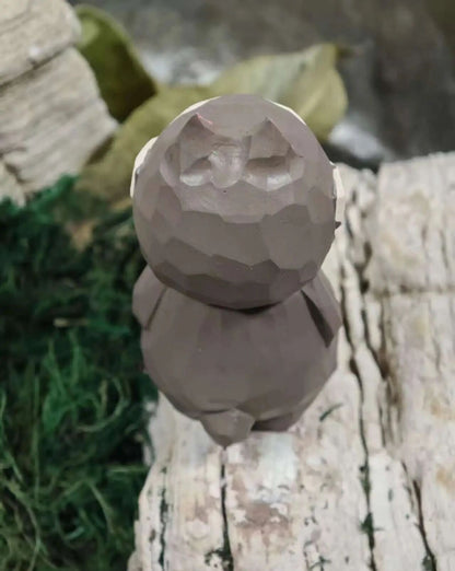A small faceted, low-poly animal figurine, likely a rabbit or hare, in a matte taupe-brown color, standing on a weathered log with blurred greenery and rocks in the background.