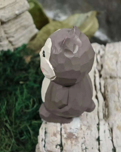 A low-poly geometric animal figurine, resembling a bear or squirrel, in shades of brown and off-white, sitting on a textured white stone surface with green moss and blurred natural elements in the background.