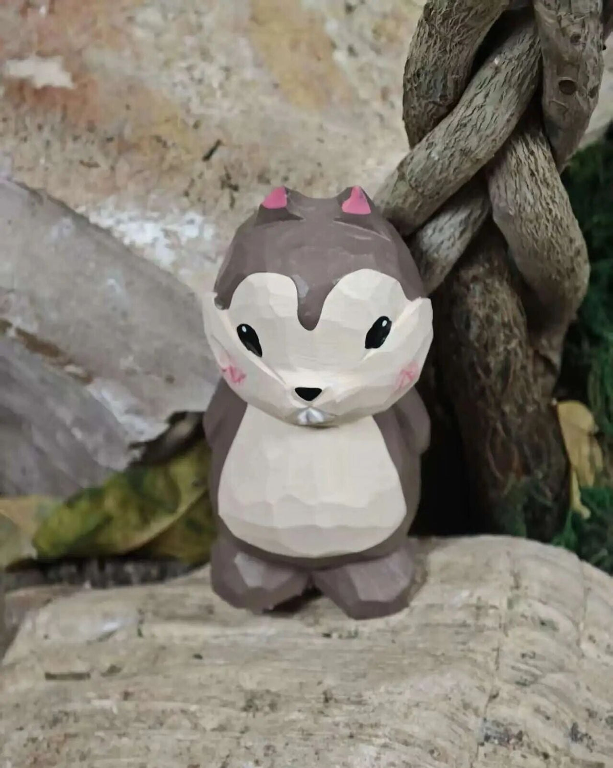 A low-poly style cartoon squirrel figurine with a brown and cream body, pink ears, and black eyes, standing on a textured stone surface next to a twisted tree branch.