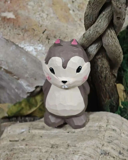 A low-poly style cartoon squirrel figurine with a brown and cream body, pink ears, and black eyes, standing on a textured stone surface next to a twisted tree branch.
