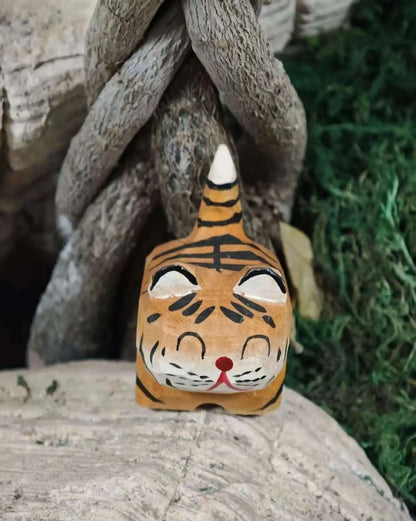 A hand-painted tiger head figurine or mask with orange, black, and white details, placed on a stone surface in front of a large twisted tree root with green grass in the background.