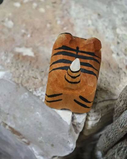 A carved wooden tiger-face bead or ornament with black painted stripes and a white tip, resting on rough natural stones.