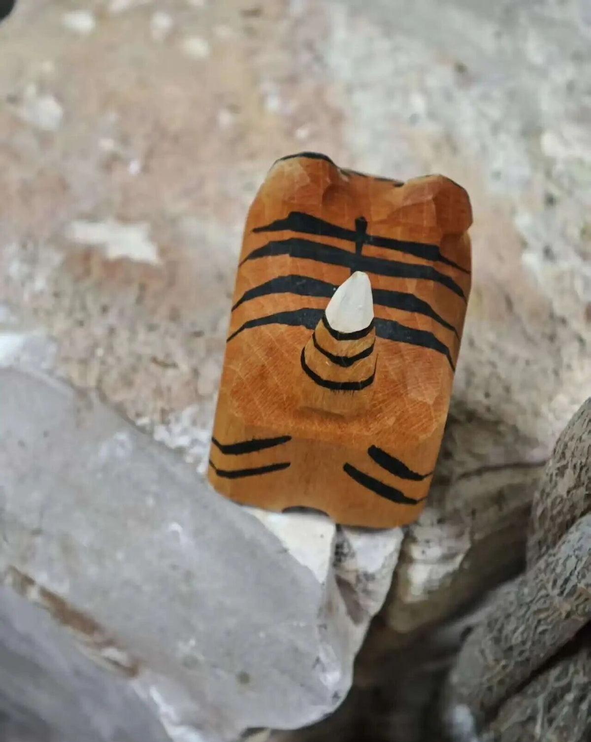 A carved wooden tiger-face bead or ornament with black painted stripes and a white tip, resting on rough natural stones.