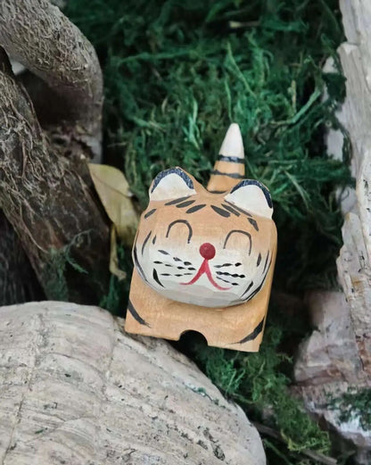 A hand-painted wooden tiger figurine with a smiling face, resting on a stone next to green moss and natural wood pieces.