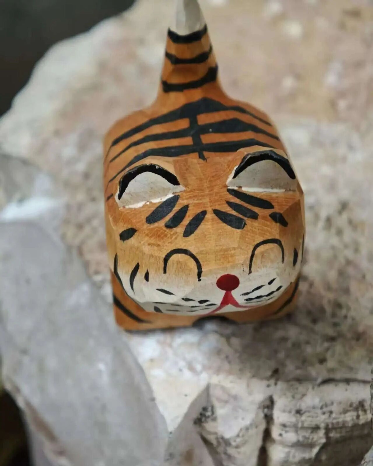 A hand-painted wooden tiger figurine with black stripes, white eyes, and a red nose, resting on a textured light gray stone surface.