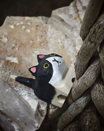 A hand-painted wooden cat-shaped desk accessory with a black and white body, pink ears, and yellow eyes, shown nestled between rustic wood and stone.