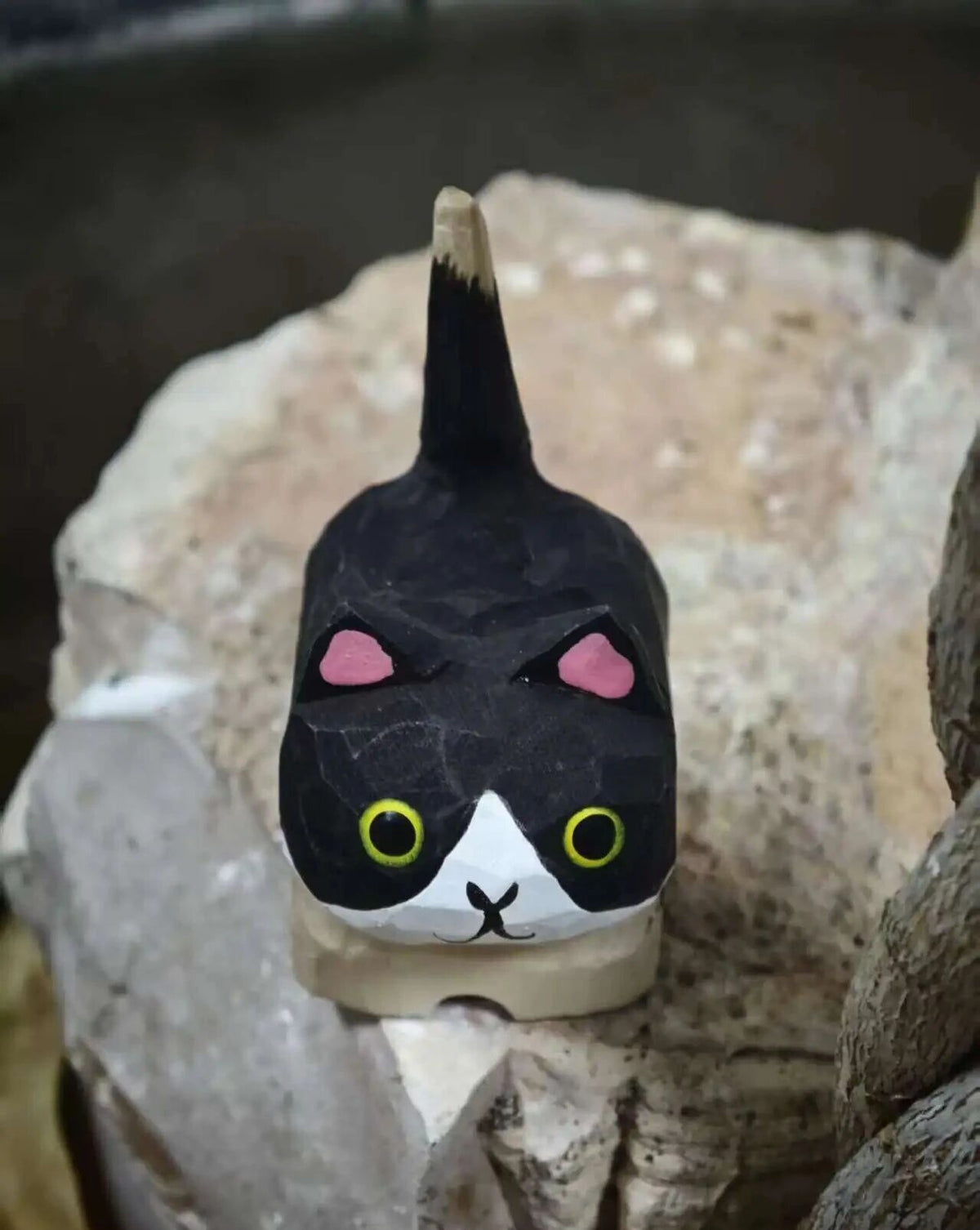 A hand-painted cat figurine with a black and white face, pink ears, and yellow eyes, resting on a textured stone surface.