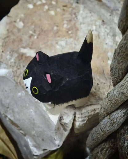 A small black-and-white cat-shaped decorative object with pink inner ears, yellow eyes, and a cream-tipped tail, photographed among rough stone and rope-like textures.