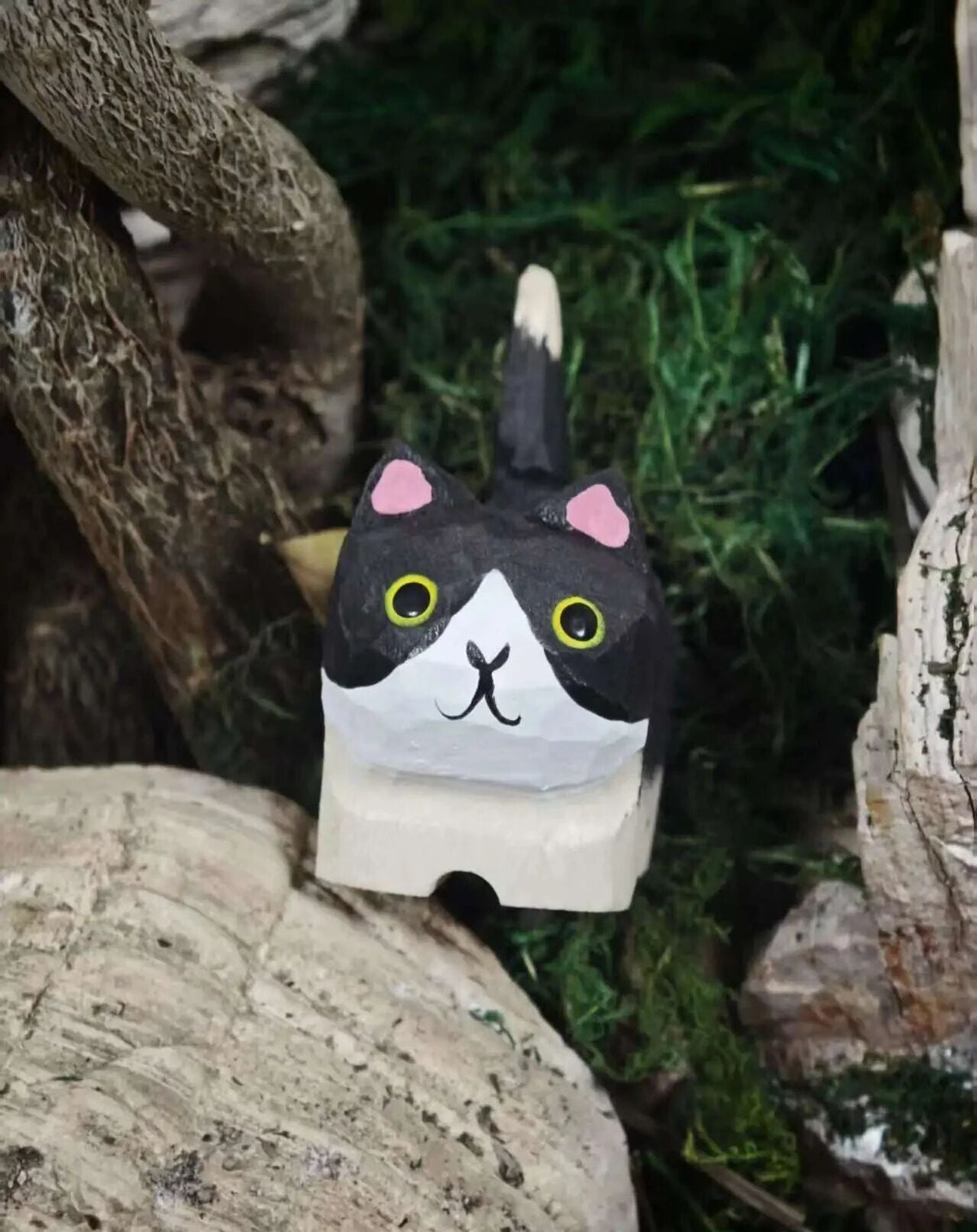 A hand-painted cat-shaped wooden block toy with a black and white face, pink ears, and yellow eyes, set against a natural background of wood and green moss.