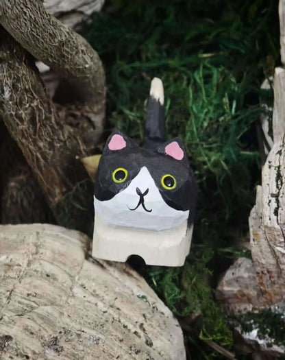 A hand-painted cat-shaped wooden block toy with a black and white face, pink ears, and yellow eyes, set against a natural background of wood and green moss.