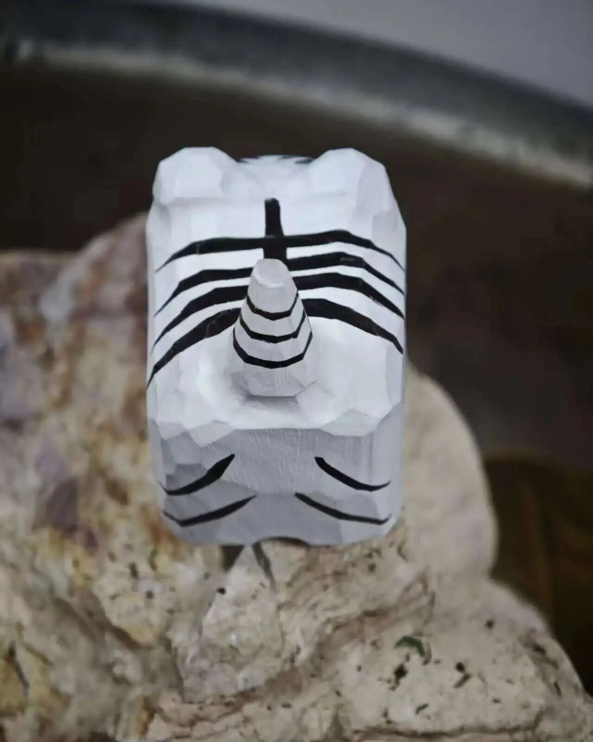 A small white geometric figurine or decorative object with black painted stripe details, standing on a textured stone surface.