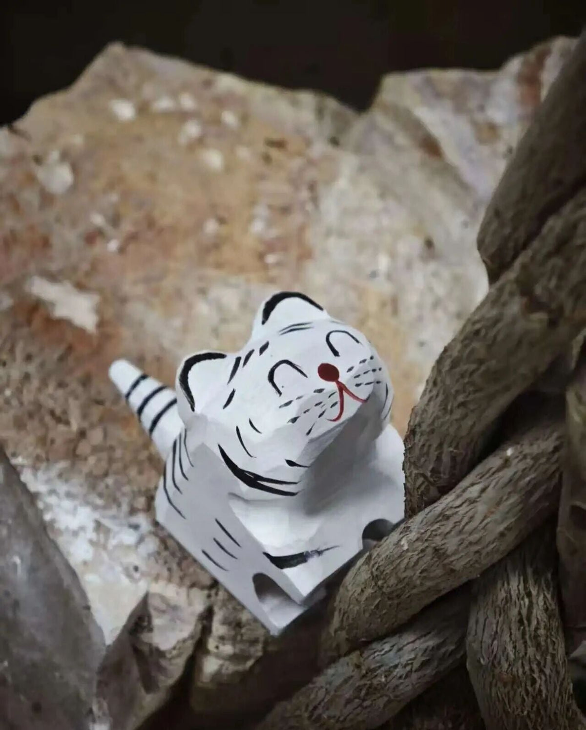 A small white carved tiger figurine with black stripes and a red nose, placed among rough stones and thick brown rope.