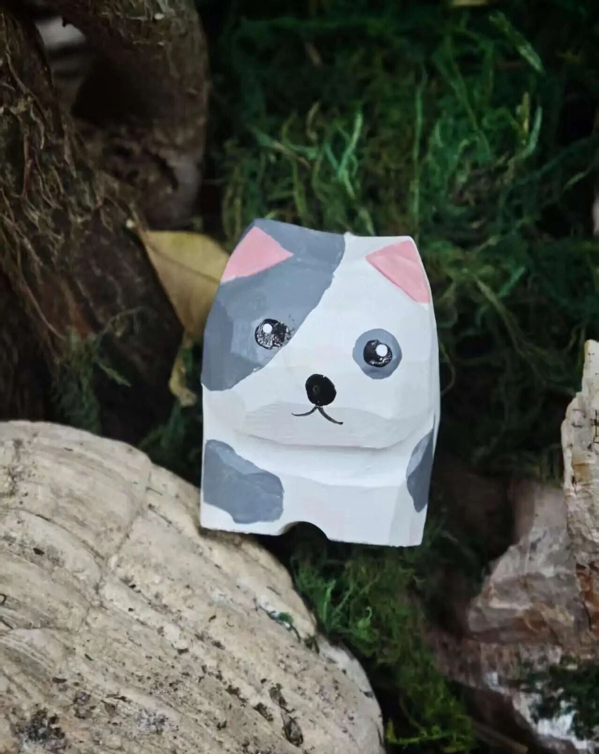 A handmade paper cat-shaped desk mat styled on a natural moss and wood surface. The cat features a white base with gray patches, pink inner ears, and simple black facial features.