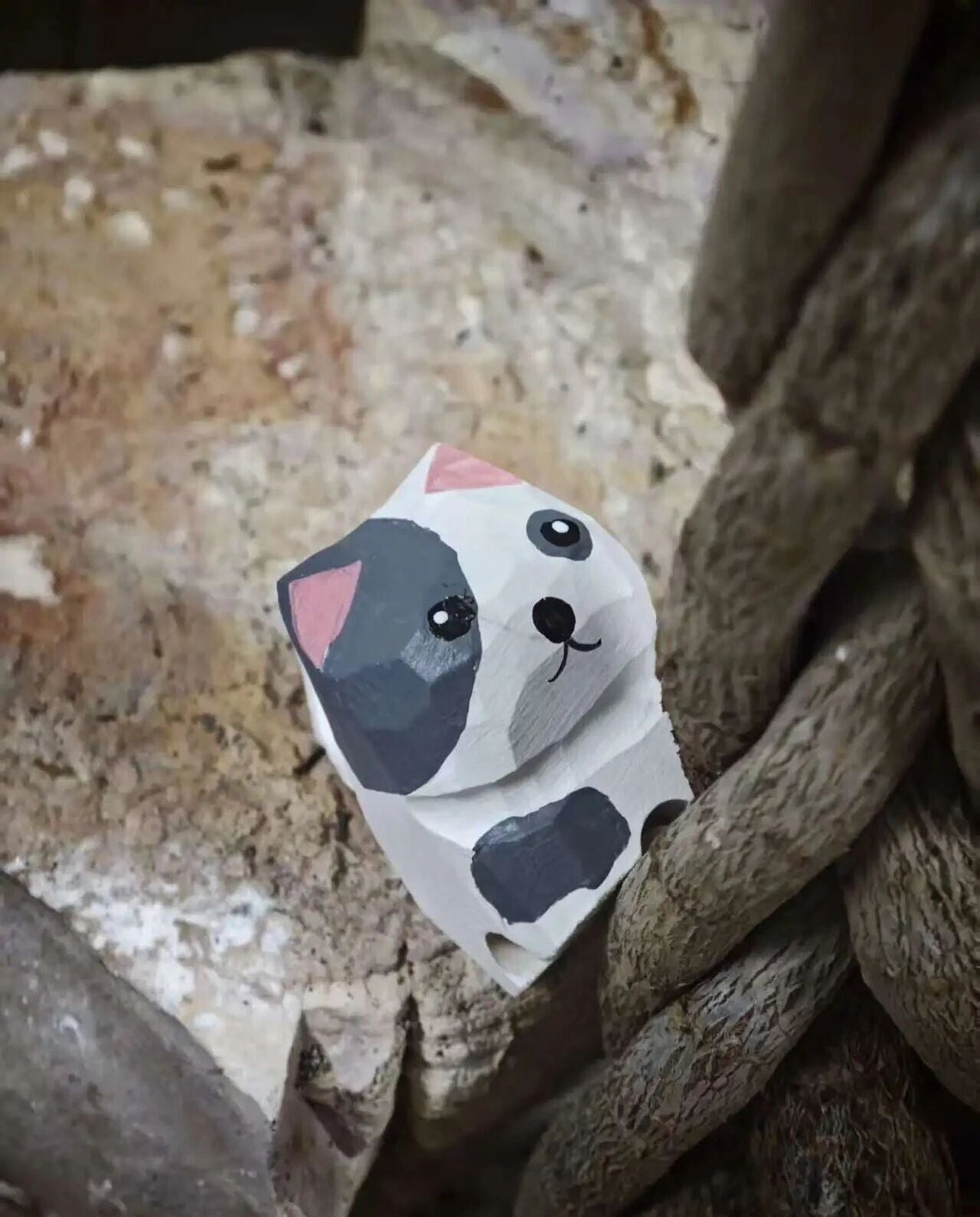 A small hand-painted wooden cat figurine with a white base, black spots, and pink ears, positioned between rustic wooden logs and stone.