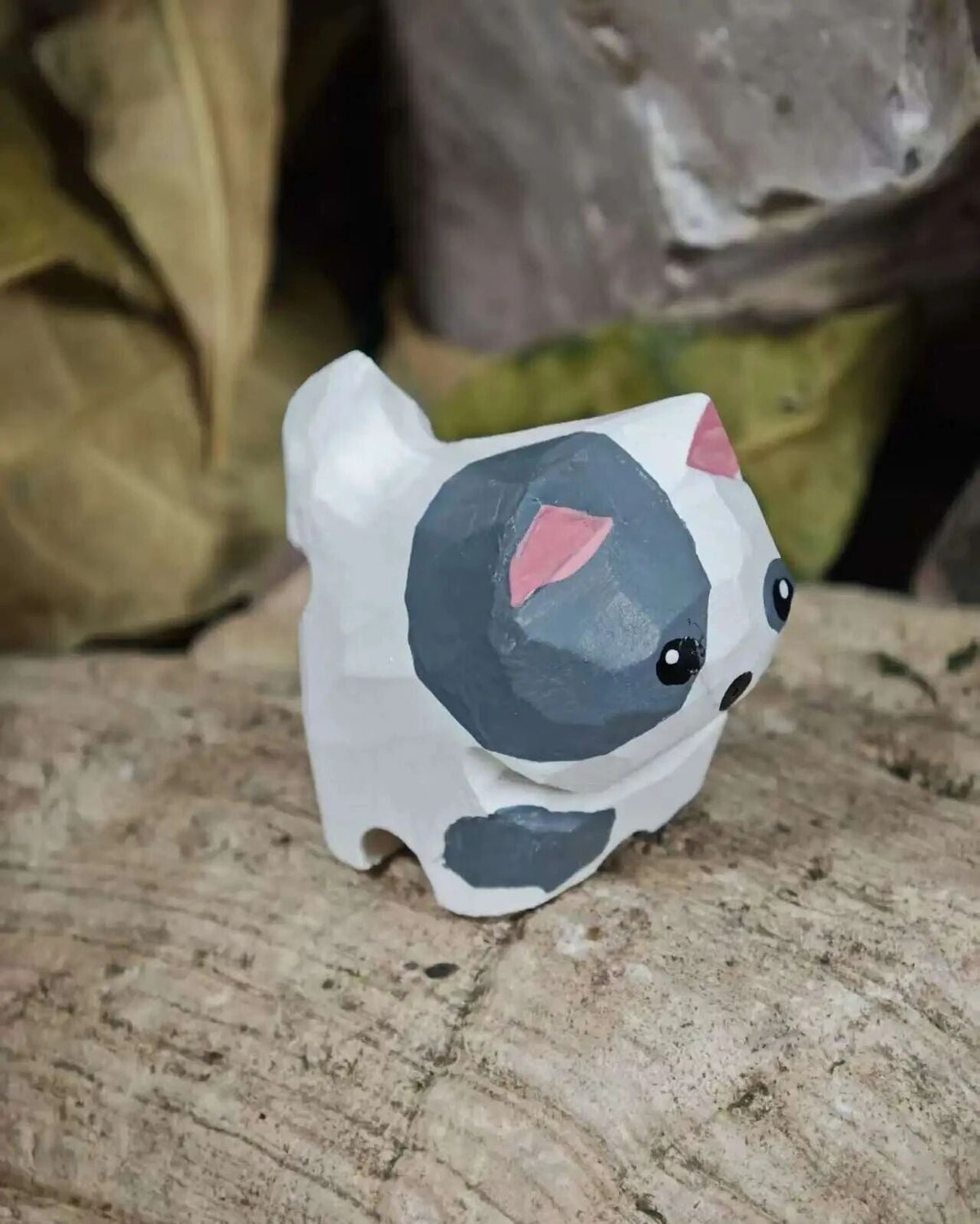 A small handcrafted wooden figurine of a cat, painted with white, dark gray, and pink, resting on a textured wooden log with a blurred natural background.