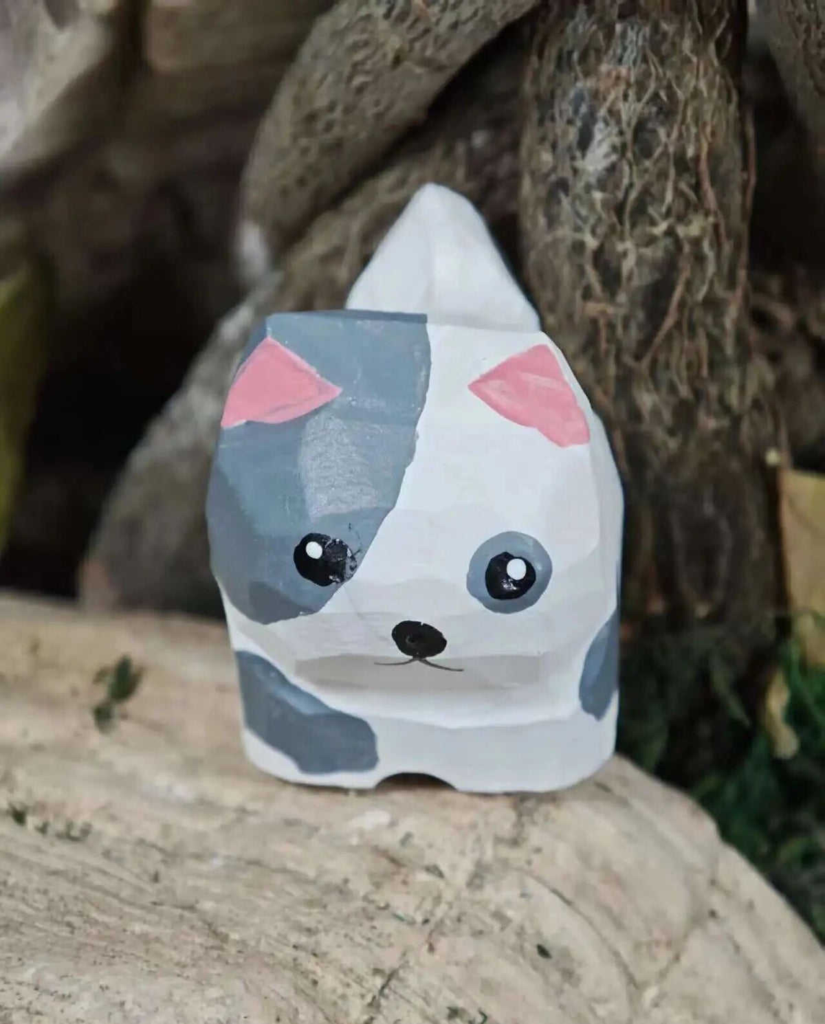 A small hand-painted ceramic figurine shaped like a cute cat head, featuring a white base with gray patches, pink inner ears, and black facial details, displayed on a stone surface outdoors.